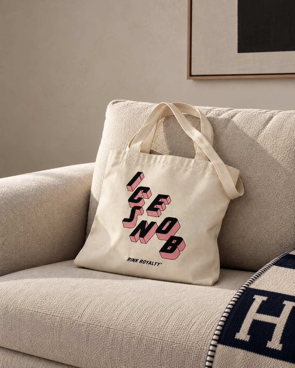An off-white canvas tote bag with the words 'ICE SNOB' in a pink and black 3D-style font, resting on a textured beige couch in a modern living room setting.